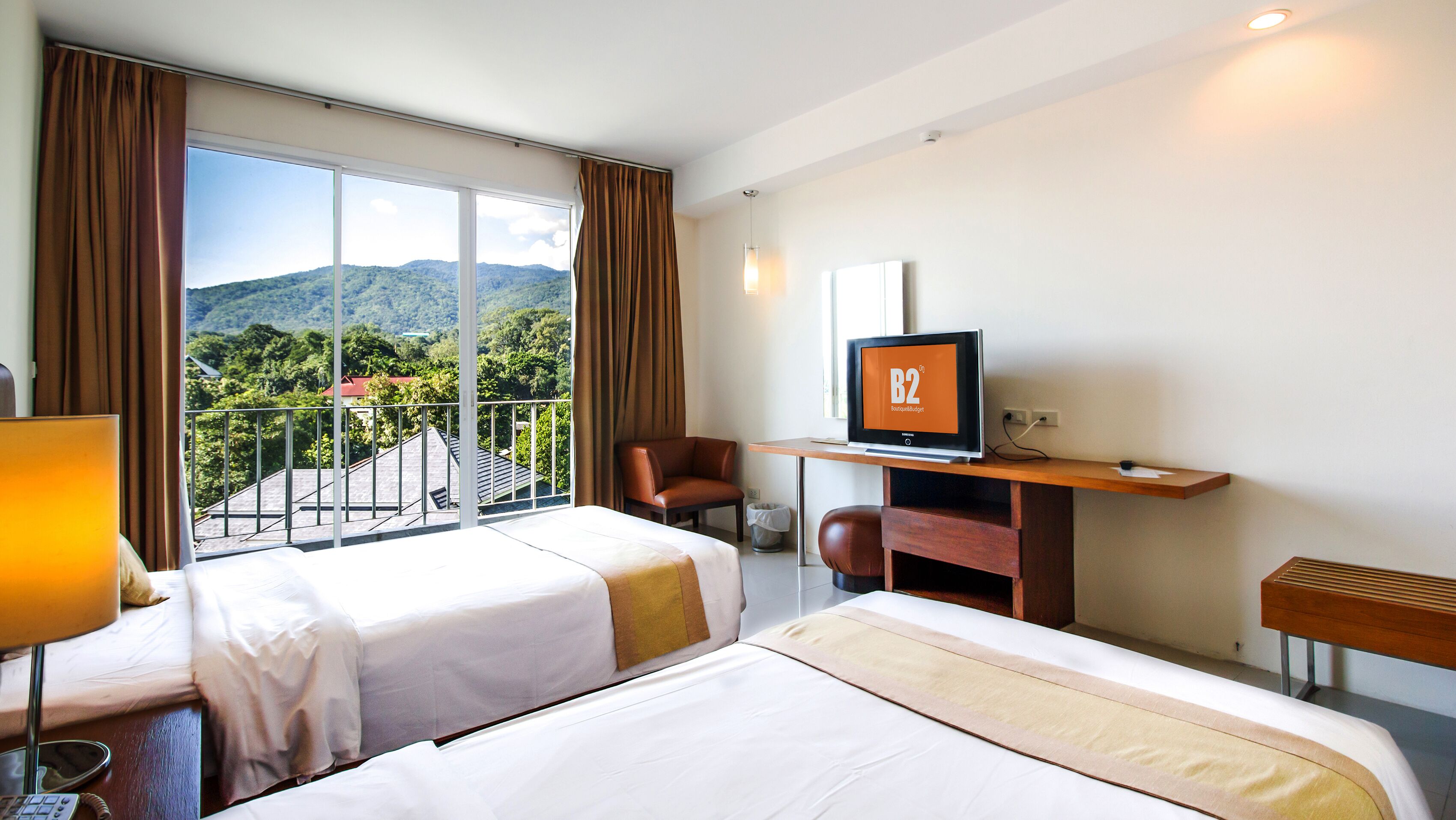 A hotel room with two beds, a desk with a TV, a chair, and a view of a mountainous landscape.