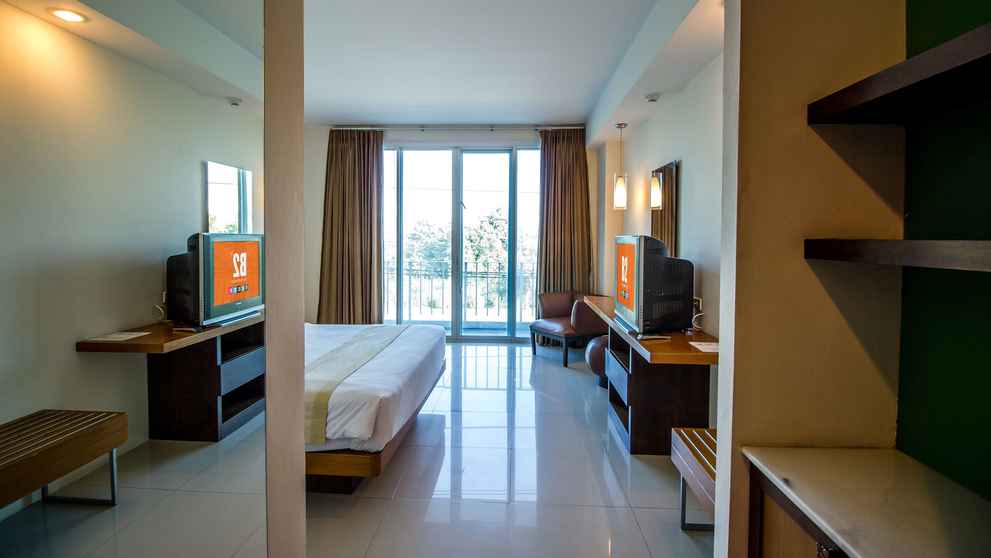 A hotel room with a bed, two TVs, a desk, a chair, and a balcony with curtains.