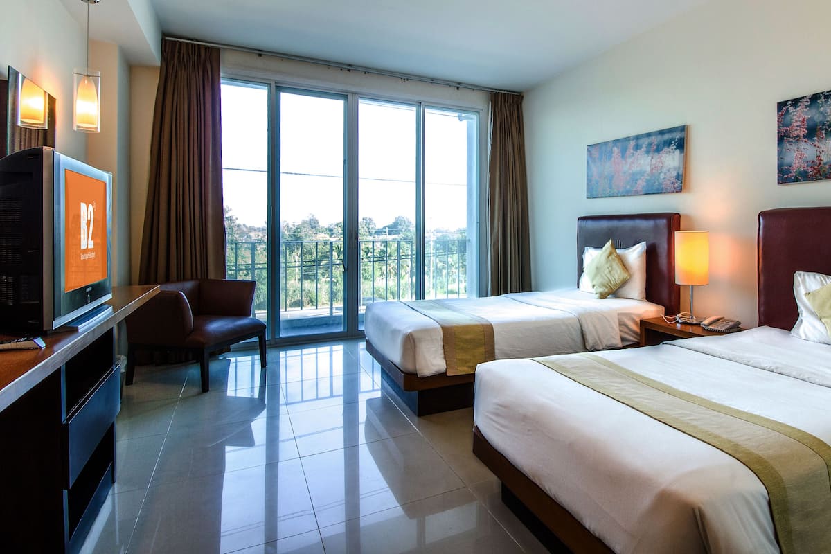 A hotel room with two beds, a desk with a TV, a chair, and a balcony with curtains.
