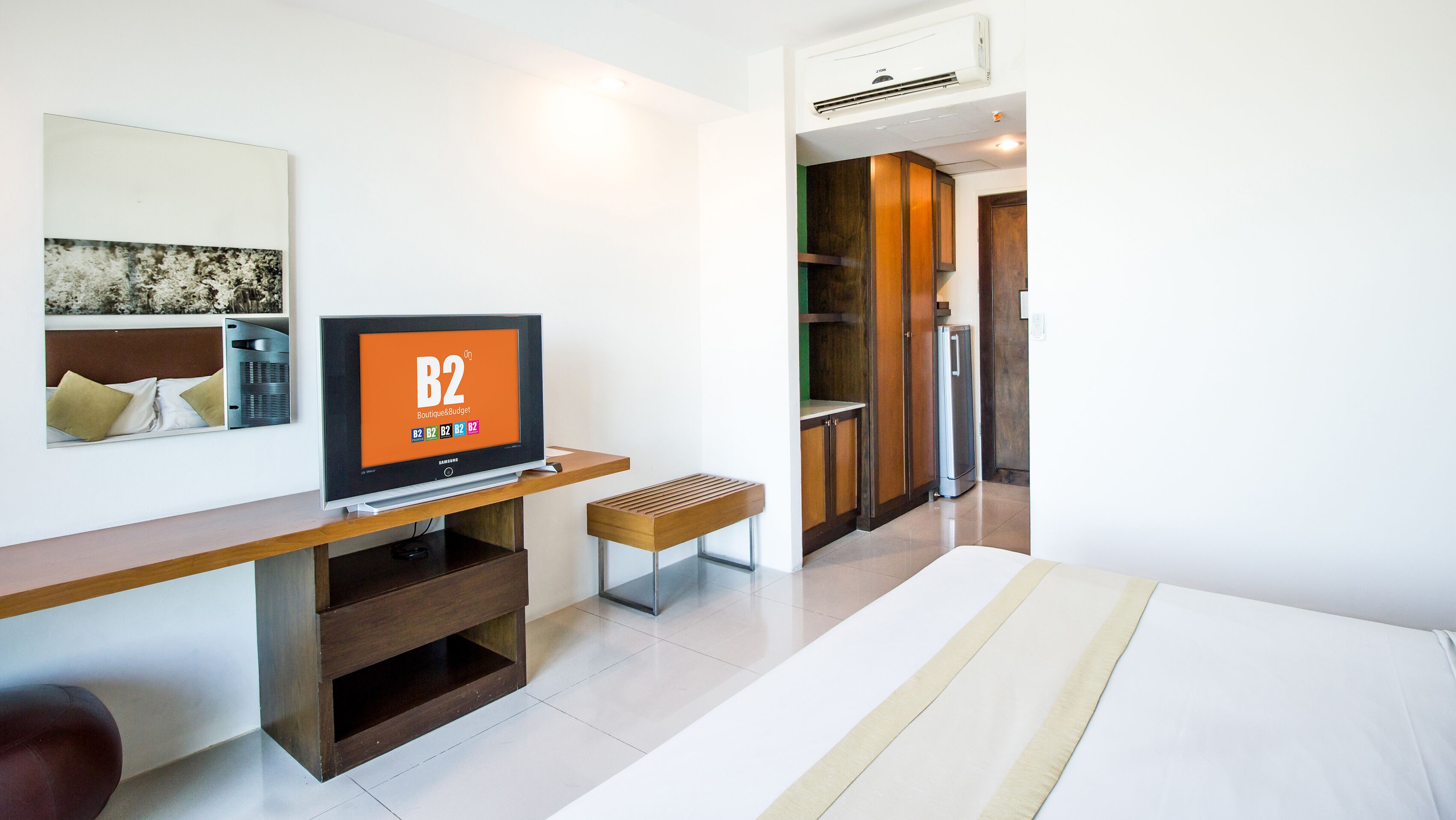 A hotel room with a TV on a wooden stand, a bed, a bench, and a kitchenette area.