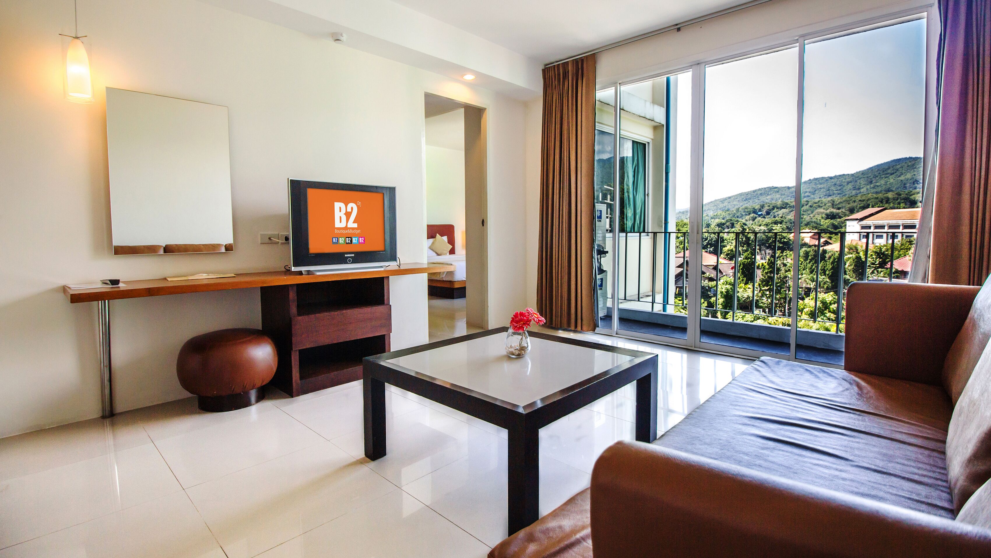 A modern hotel room with a large TV, a sofa, a coffee table, and a balcony with a view of the hills.