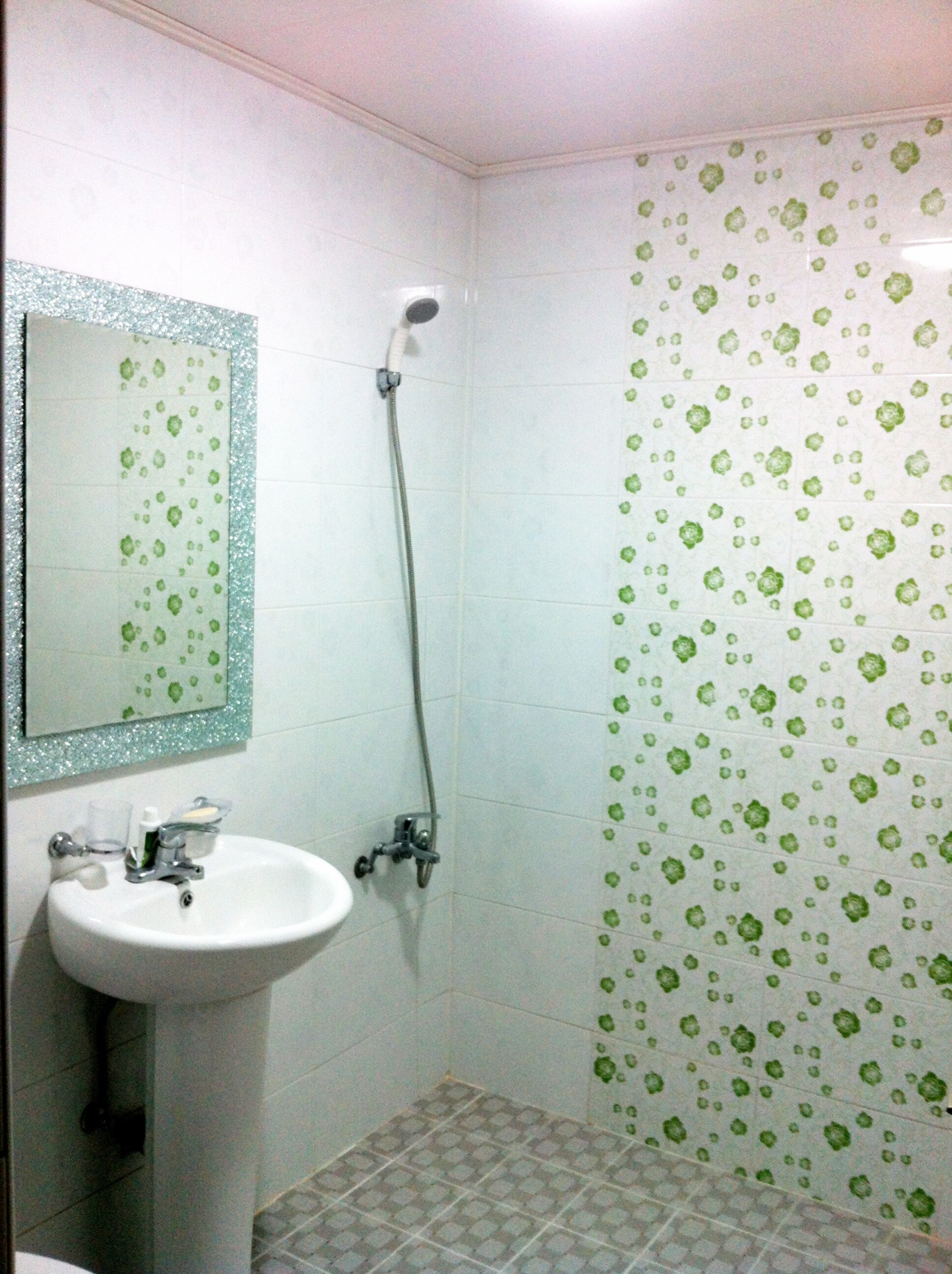 Un baño con un lavamanos blanco, un espejo con marco decorativo, una ducha con diseño de azulejos florales verdes y suelo de baldosas.