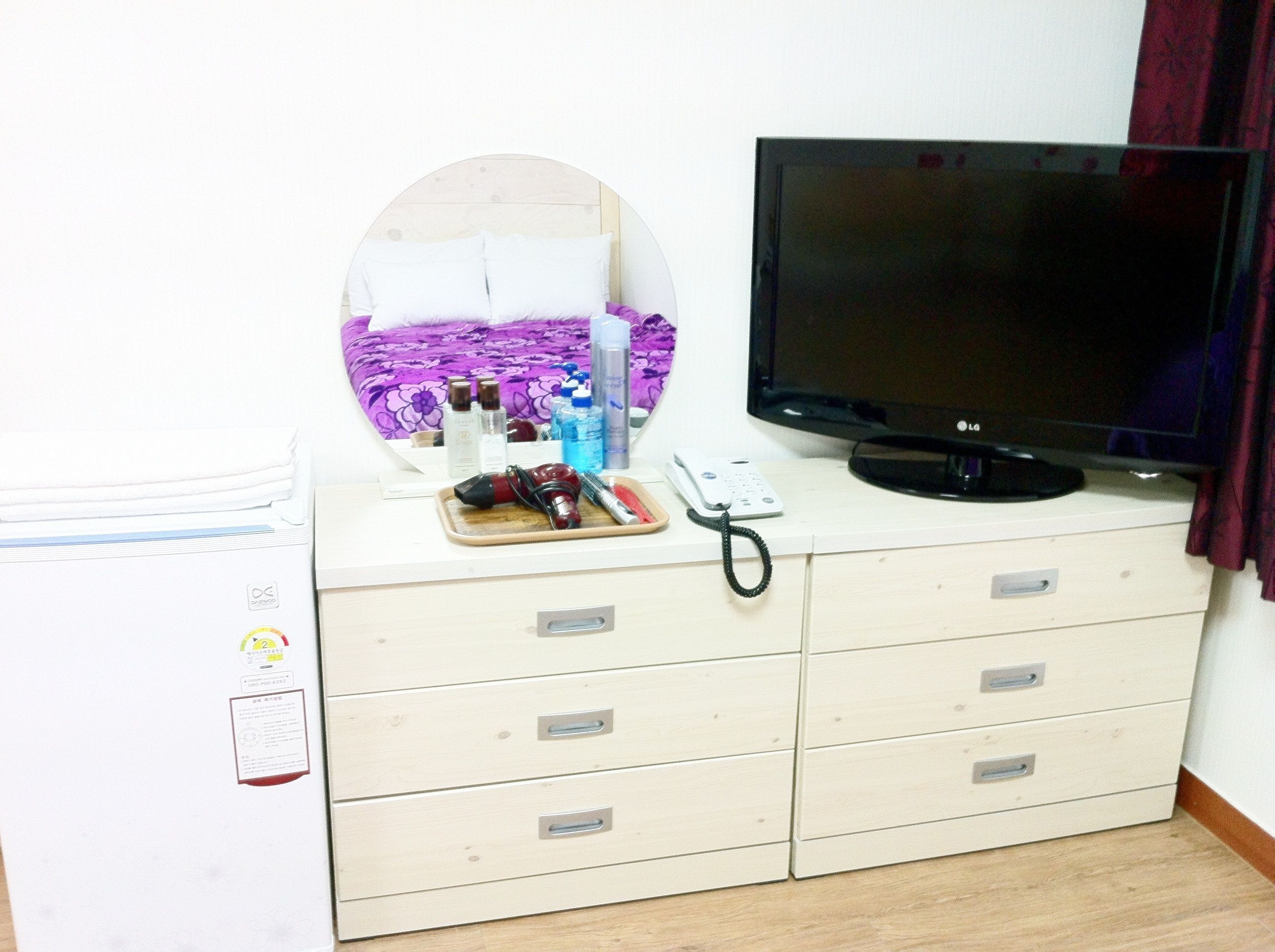 Ein Zimmer mit einem Bett, einem Fernseher, einer Kommode mit Schubladen, einem Spiegel und einem Nachttisch mit Pflegeartikeln.
