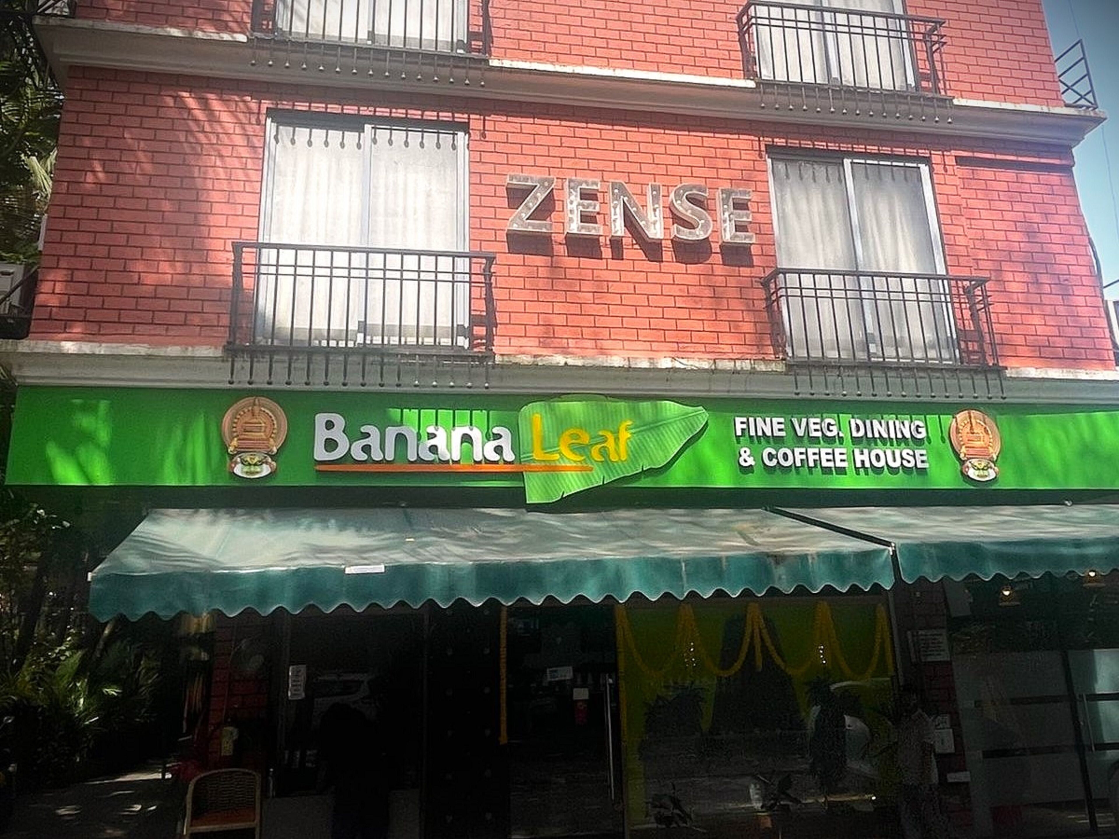 A building with a red brick facade and a green awning. The awning has the text "Banana Leaf" and "Fine Veg Dining & Coffee House".