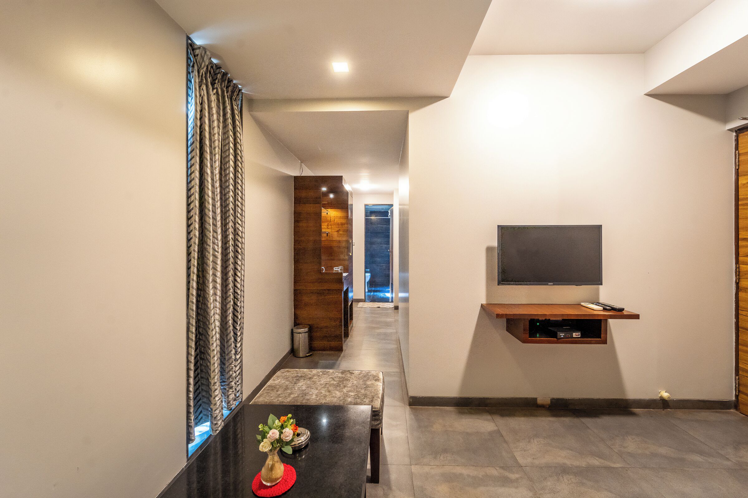 A modern hallway with a flat-screen TV mounted on a wooden shelf, a small table with a vase, and a staircase leading to another room.