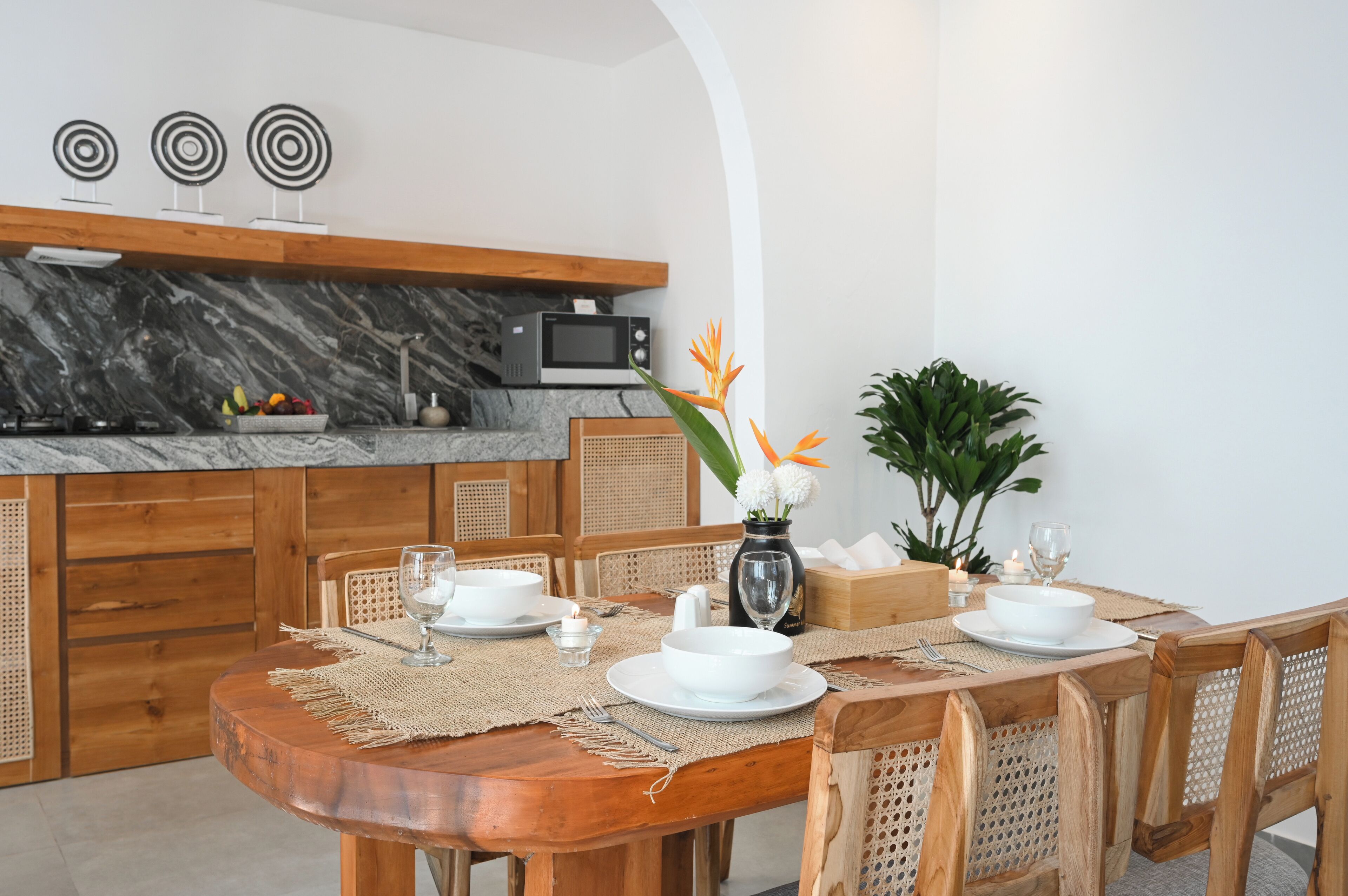 Un coin repas avec une table en bois dressée pour quatre personnes, entourée de chaises en osier. On aperçoit à l’arrière-plan une cuisine dotée d’un plan de travail en marbre et d’un four à micro-ondes.