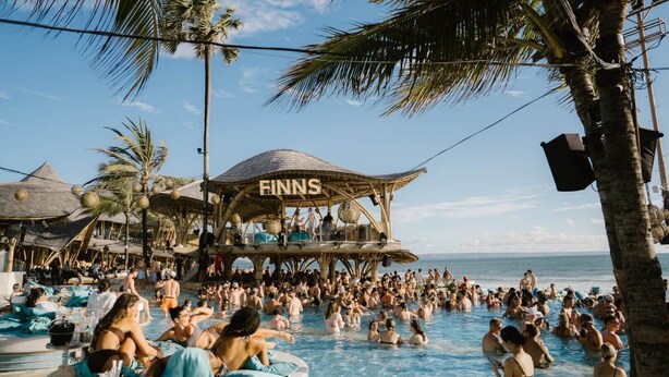 Uma área de piscina movimentada com uma estrutura de madeira identificada como 'FINNS' e palmeiras.