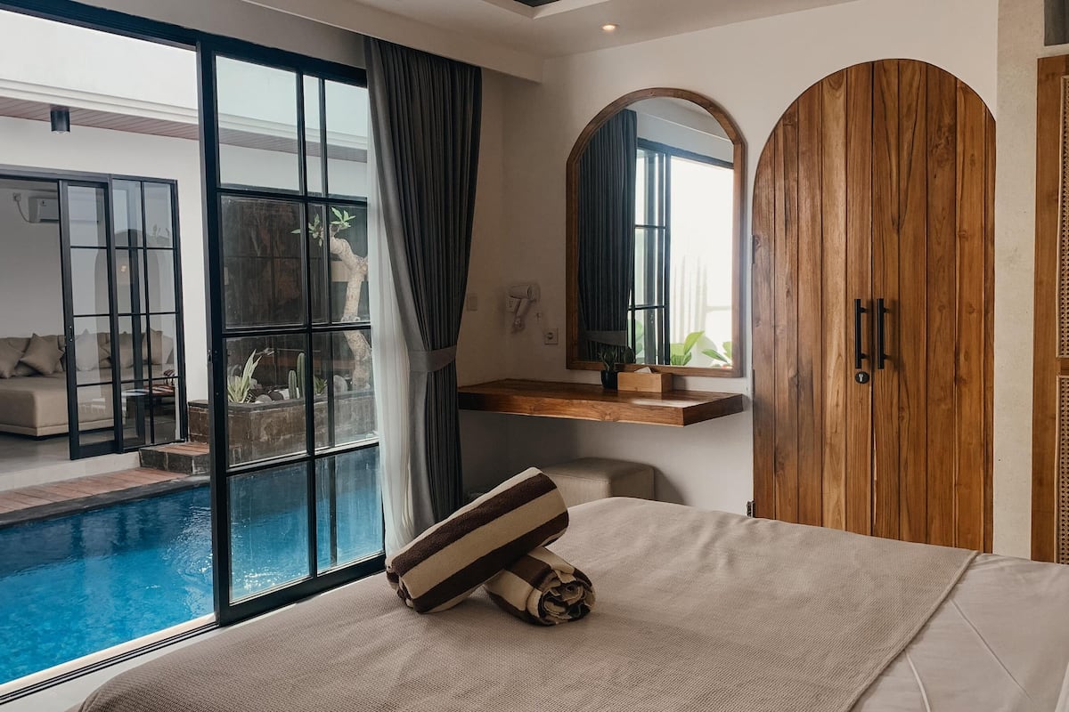 Een moderne slaapkamer met een groot glazen schuifdeur naar een zwembad, een houten garderobe en een nachtkastje met een spiegel.