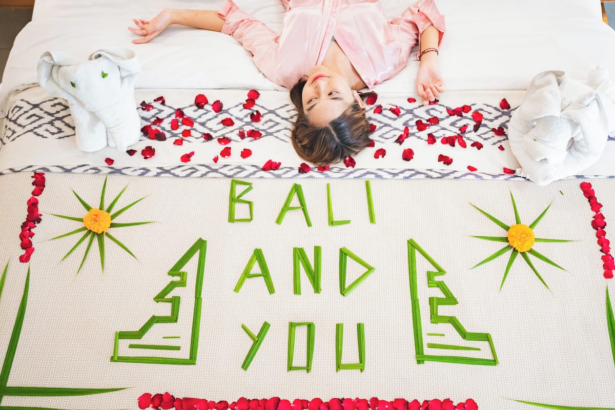 Een bed met een deken waarop 'BALI AND YOU' staat, omringd door rode bloemen en twee handdoeken gevouwen in de vorm van olifanten.