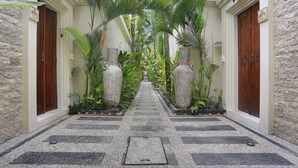 Um corredor com chão quadriculado, ladeado por paredes de pedra e portas de madeira, conduz a uma área central com plantas e um caminho.