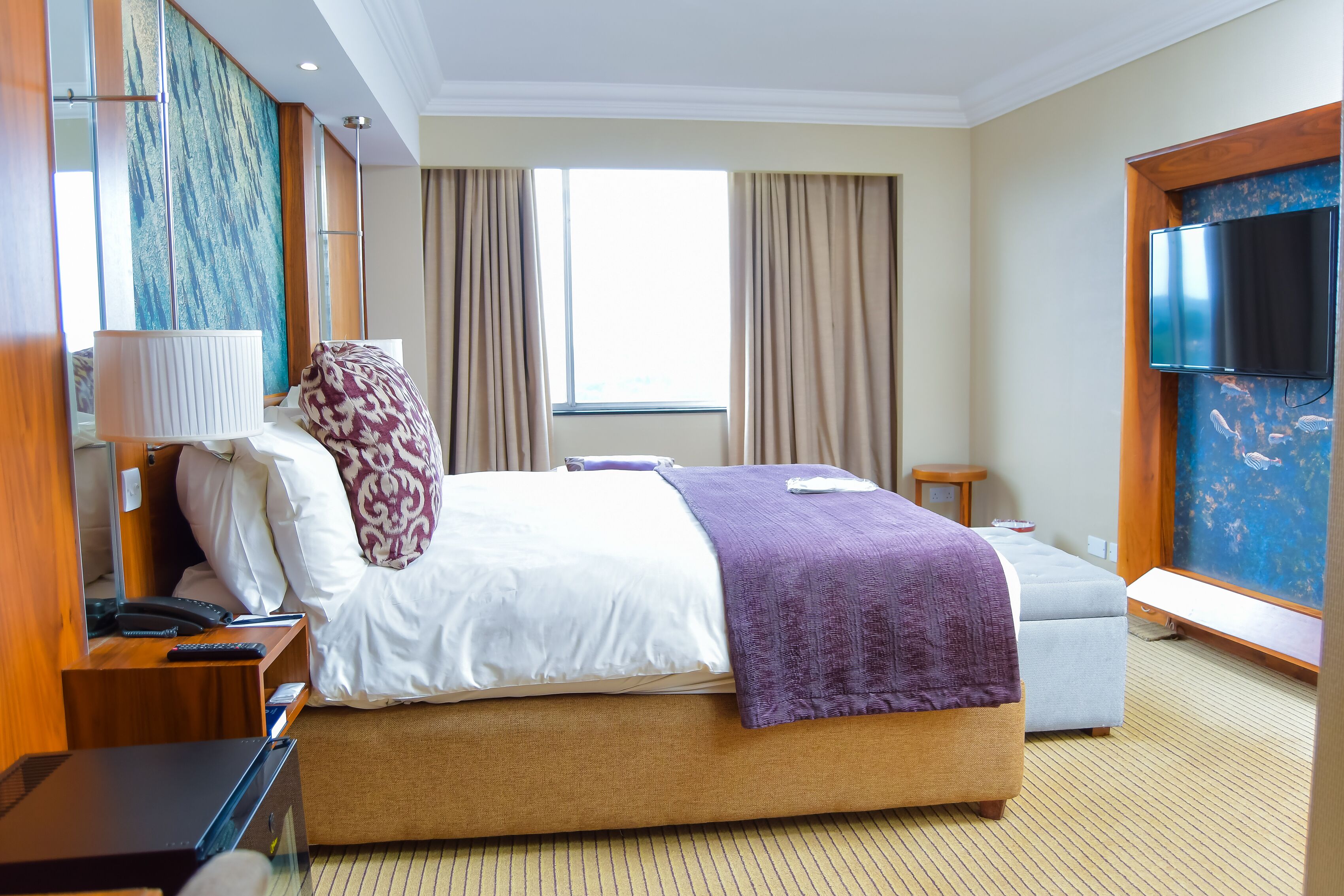 Een hotelkamer met een groot bed, een nachtkastje met een lamp, een aan de muur bevestigde televisie en een raam met gordijnen.