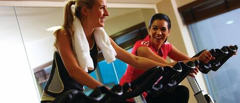 Two people on stationary bikes in a gym with mirrored walls and a window.