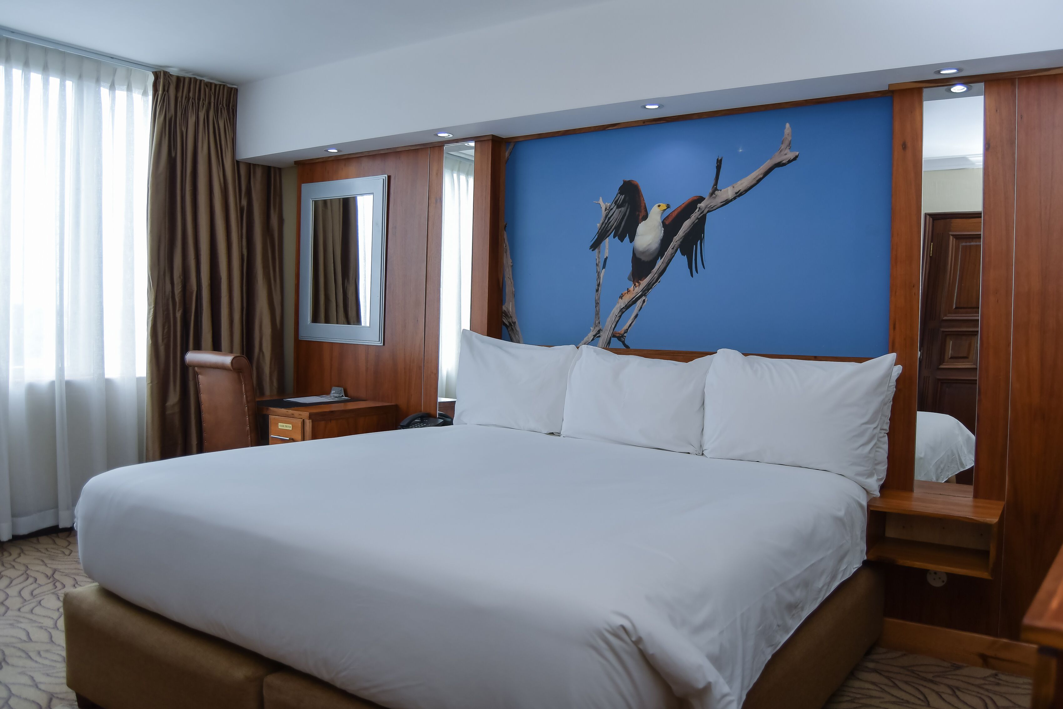 A hotel room with a large bed, a wooden nightstand, a desk, and a wall-mounted mirror.