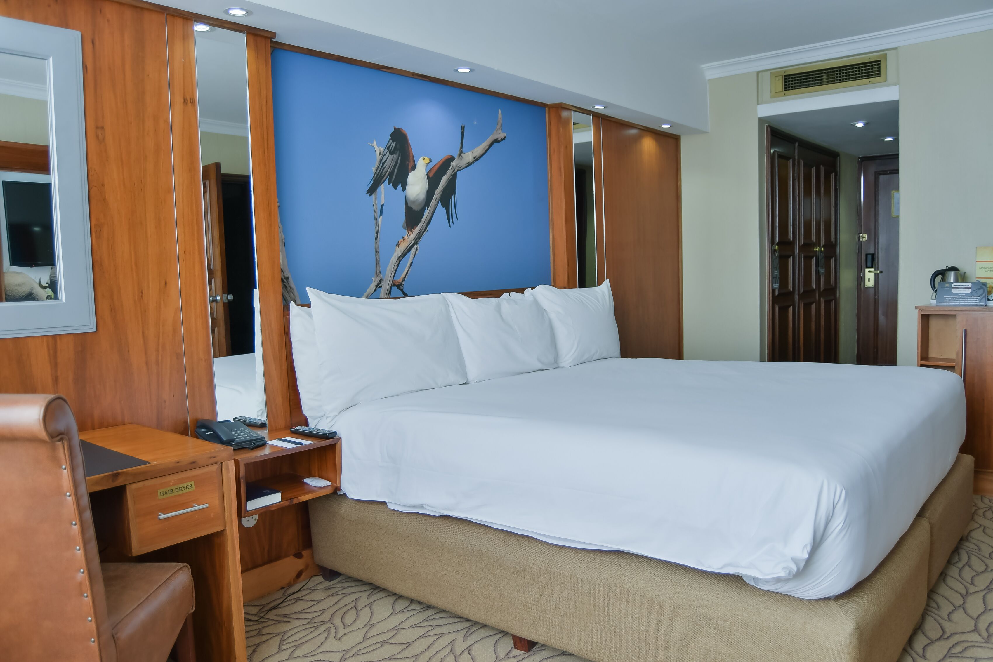 A hotel room with a large bed, wooden furniture, a desk with a hair dryer, and a wall-mounted painting of a bird.