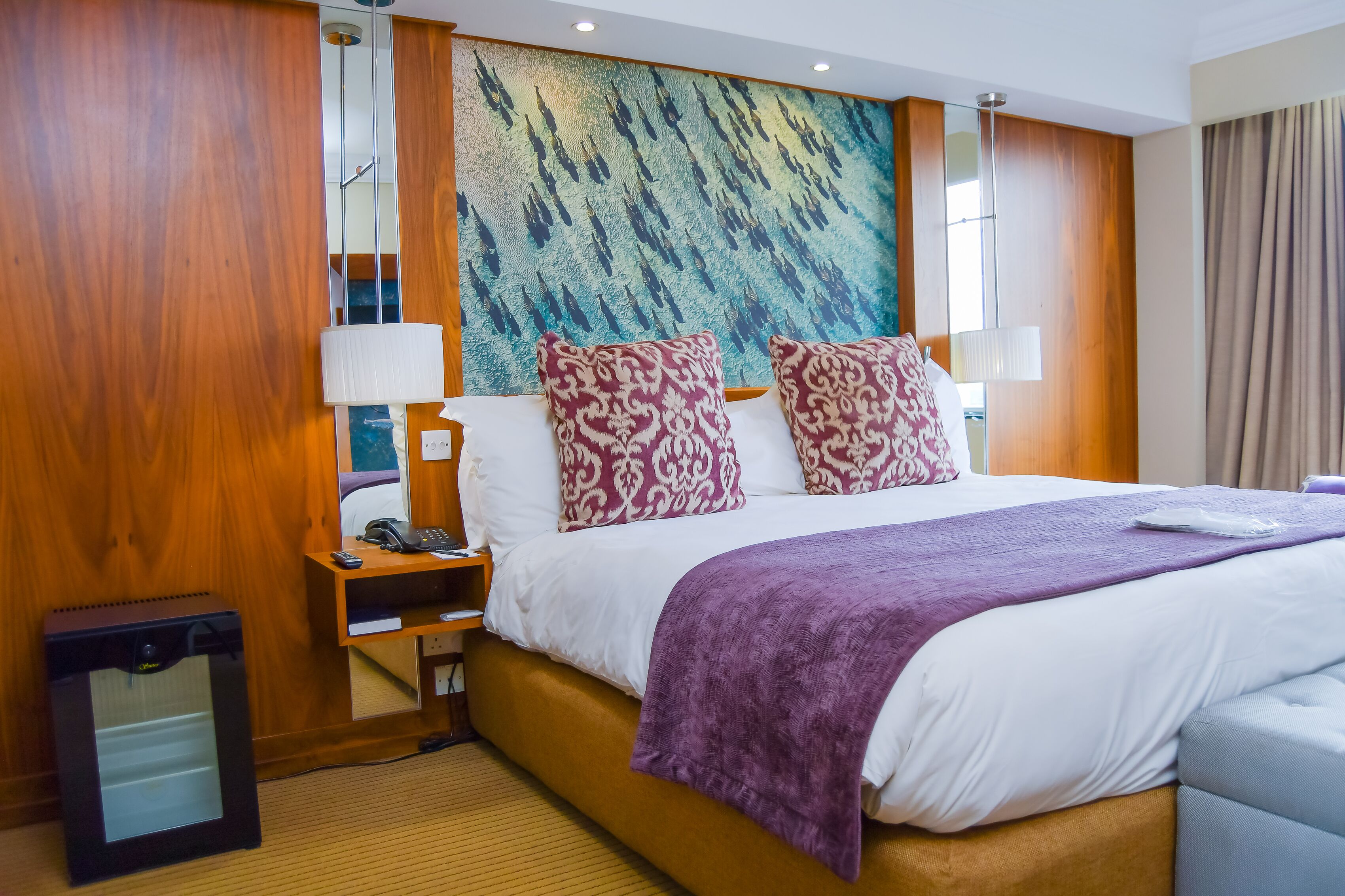 A hotel room with a large bed, wooden paneling, a large abstract painting, and a desk with a lamp.