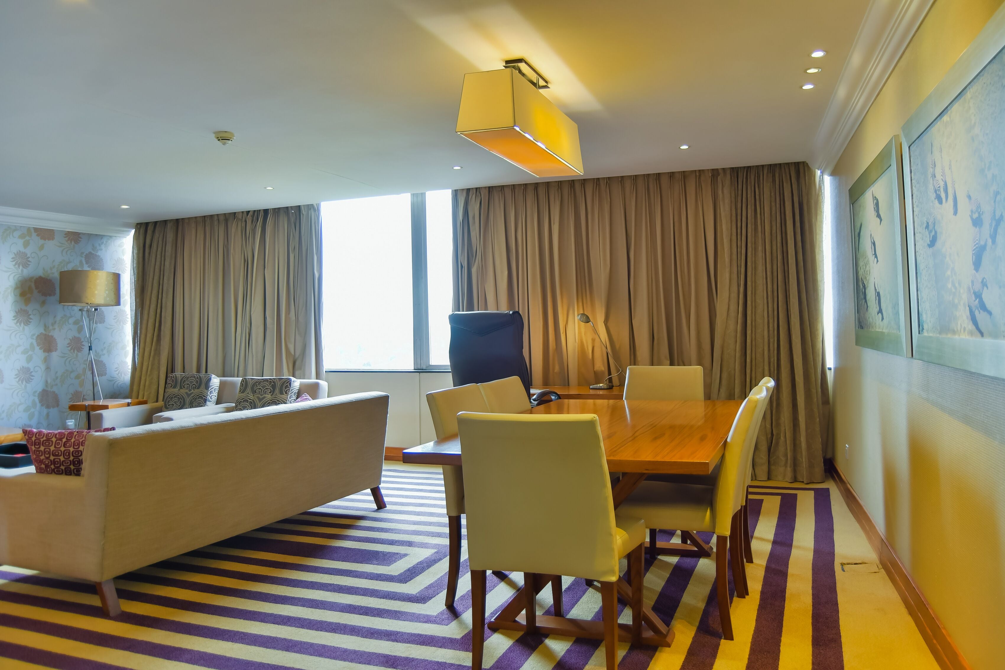 A modern hotel room with a sofa, dining table, chairs, a lamp, and a large window with curtains.