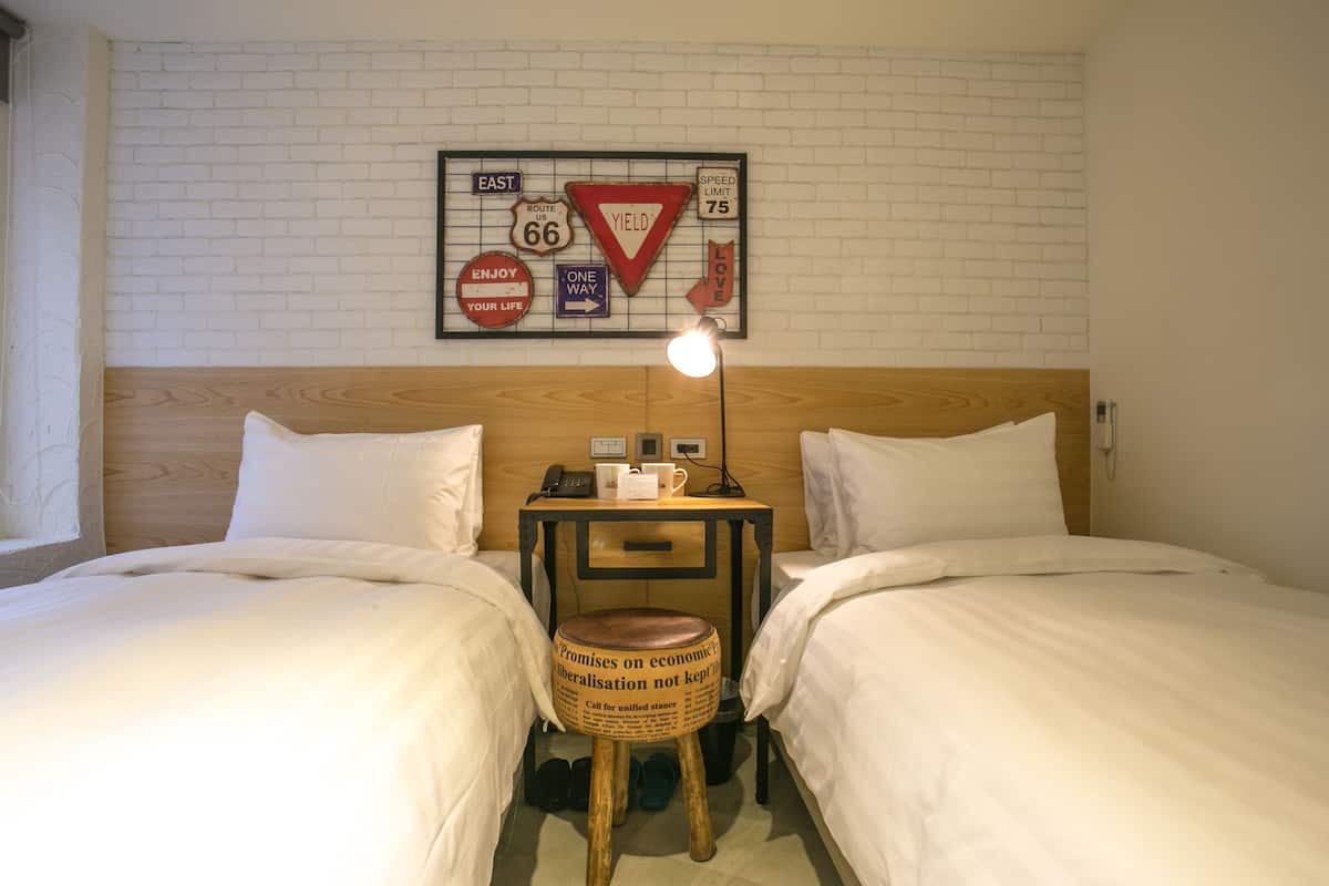 A hotel room with two beds, a wooden headboard, a small table with a lamp, and a wall decoration featuring road signs.
