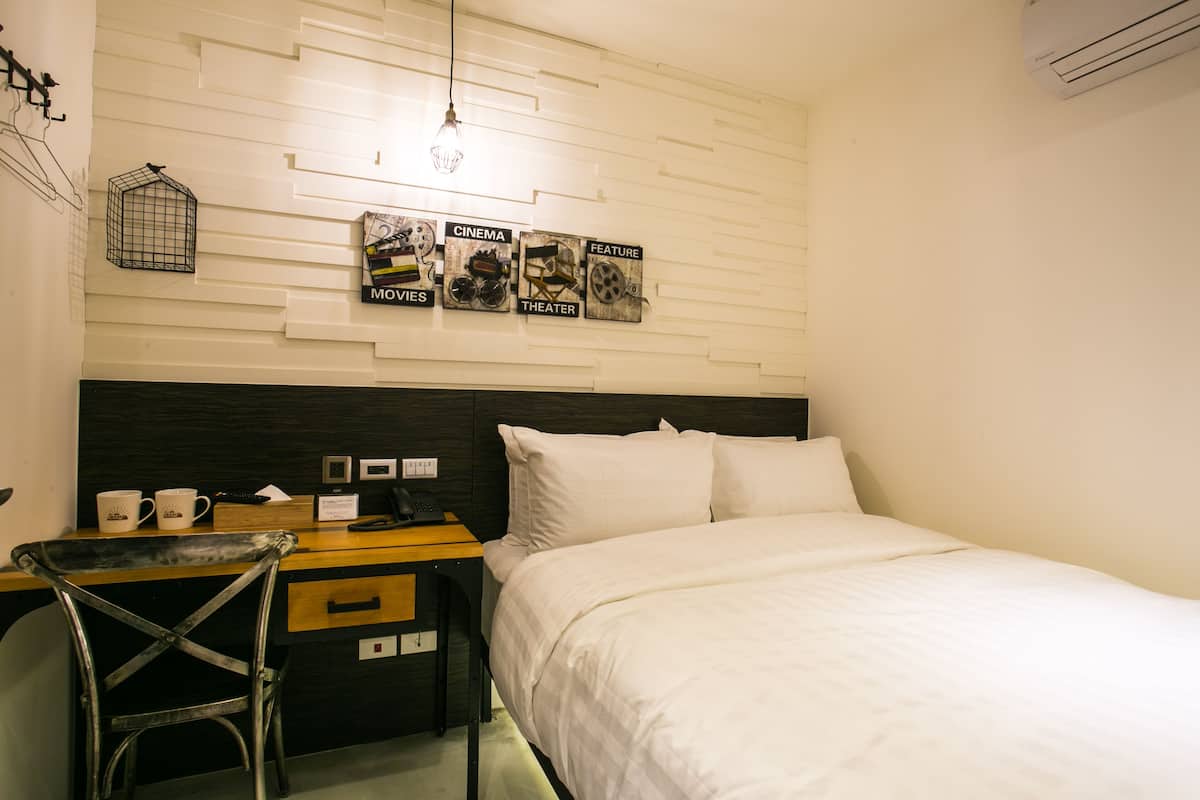 A hotel room with a bed, a desk with a chair, a wall-mounted light fixture, and a decorative wall panel with movie-related items.