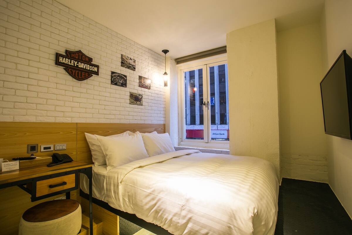 A hotel room with a white brick wall, a Harley-Davidson sign, a bed with white bedding, a wooden headboard, a desk with a telephone, and a window with a view of a building.