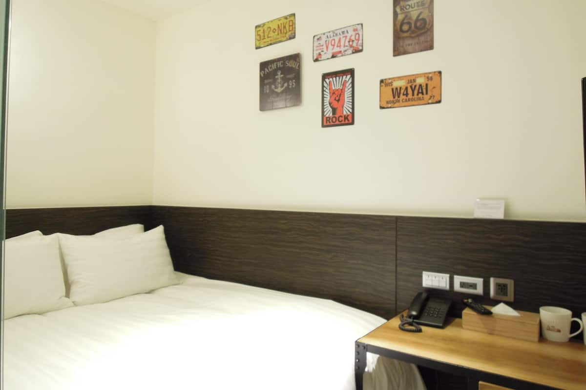 A hotel room with a bed, a bedside table with a phone and a mug, and a wall with various vintage route signs.