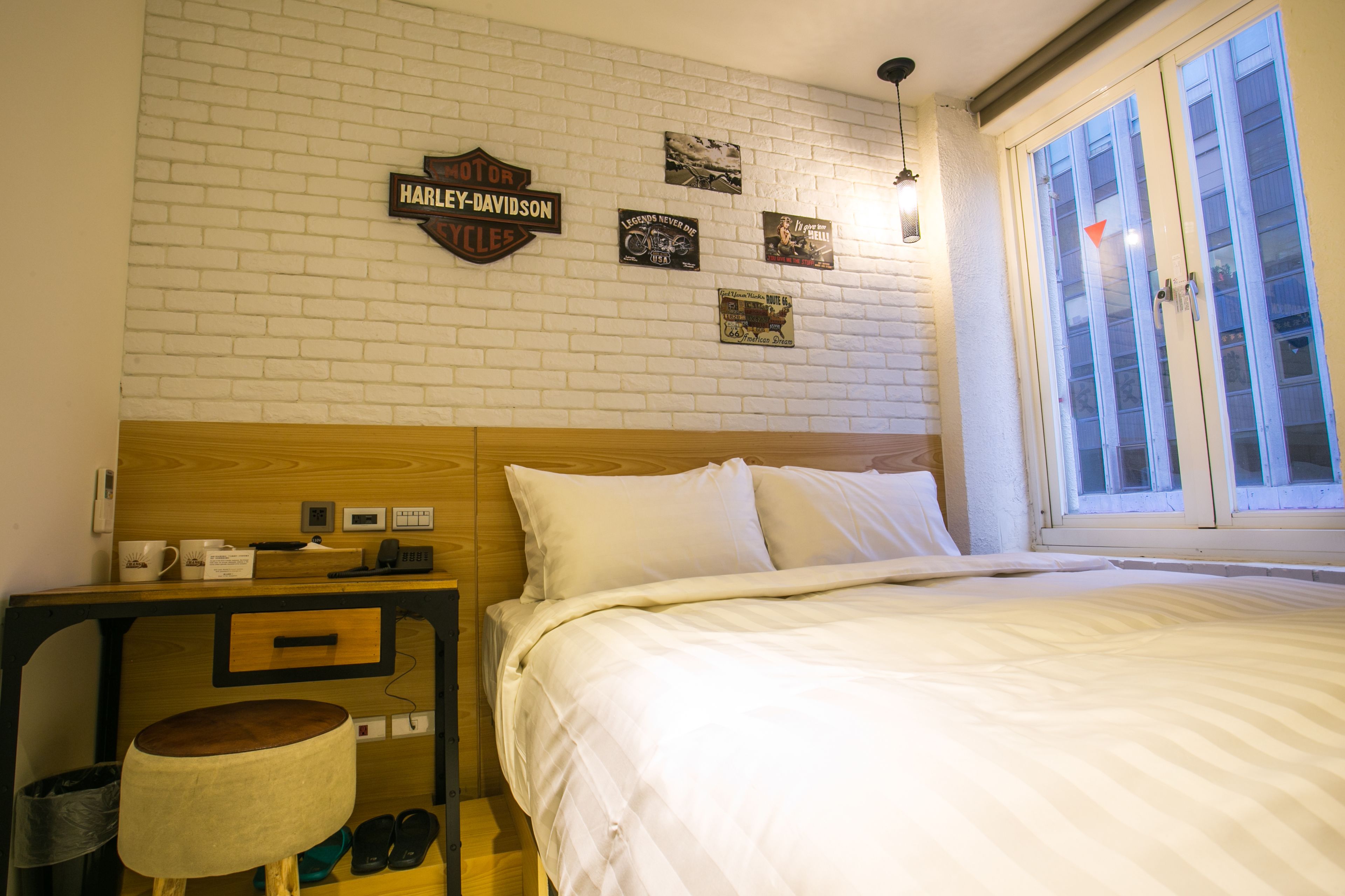 A hotel room with a bed, a nightstand, a lamp, a wooden headboard, and a window with blinds.