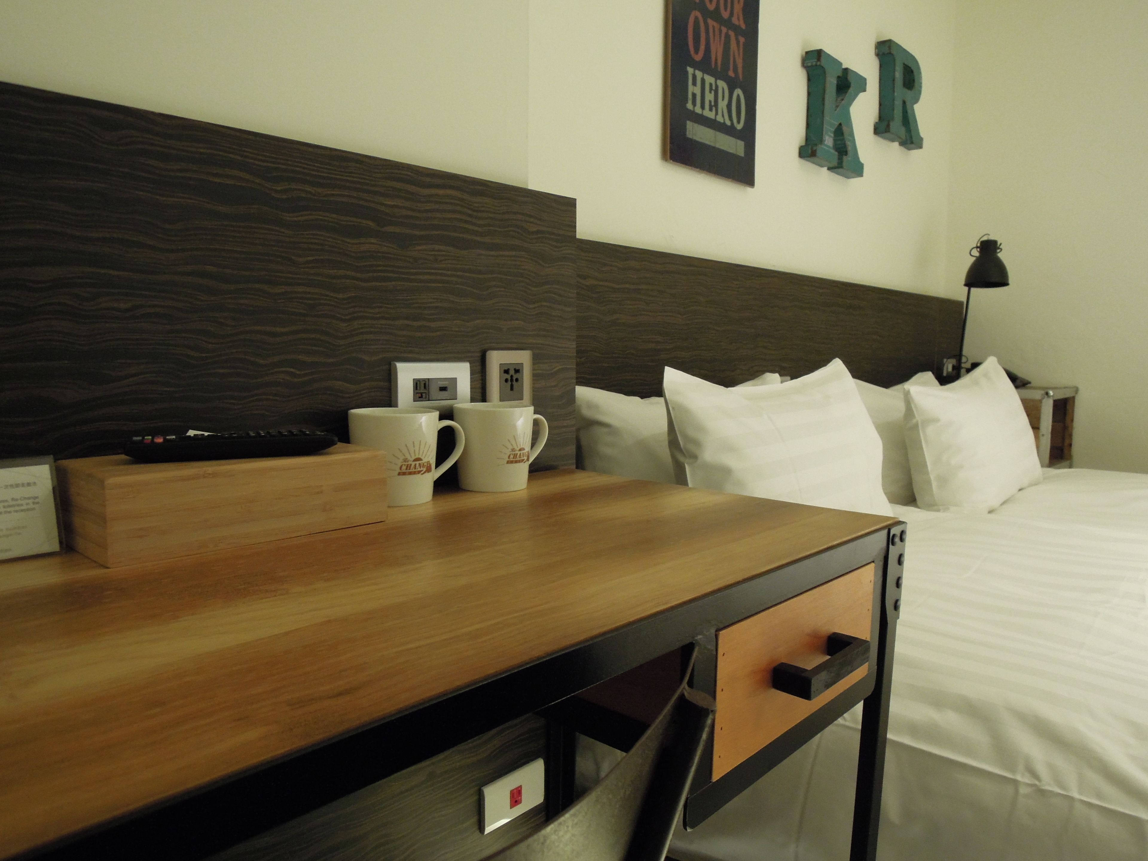 Quarto de hotel com mesa de cabeceira de madeira, duas canecas brancas, um controle remoto, tomada na parede, abajur e cama com roupa de cama branca.