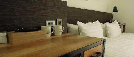Quarto de hotel com mesa de cabeceira de madeira, duas canecas brancas, um controle remoto, tomada na parede, abajur e cama com roupa de cama branca.