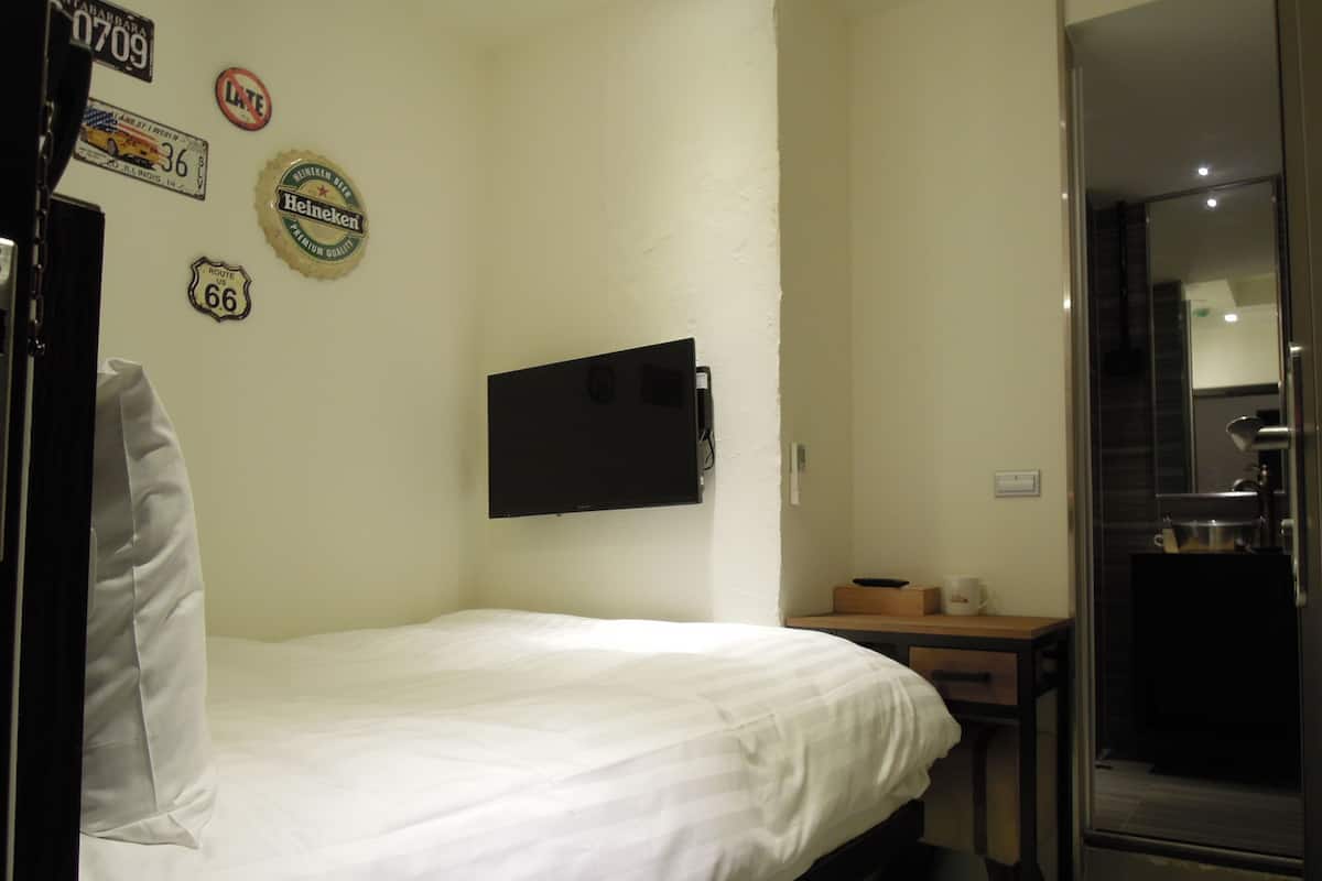 A small, clean room with a single bed, a mounted television, and a small table with a cup and a box.