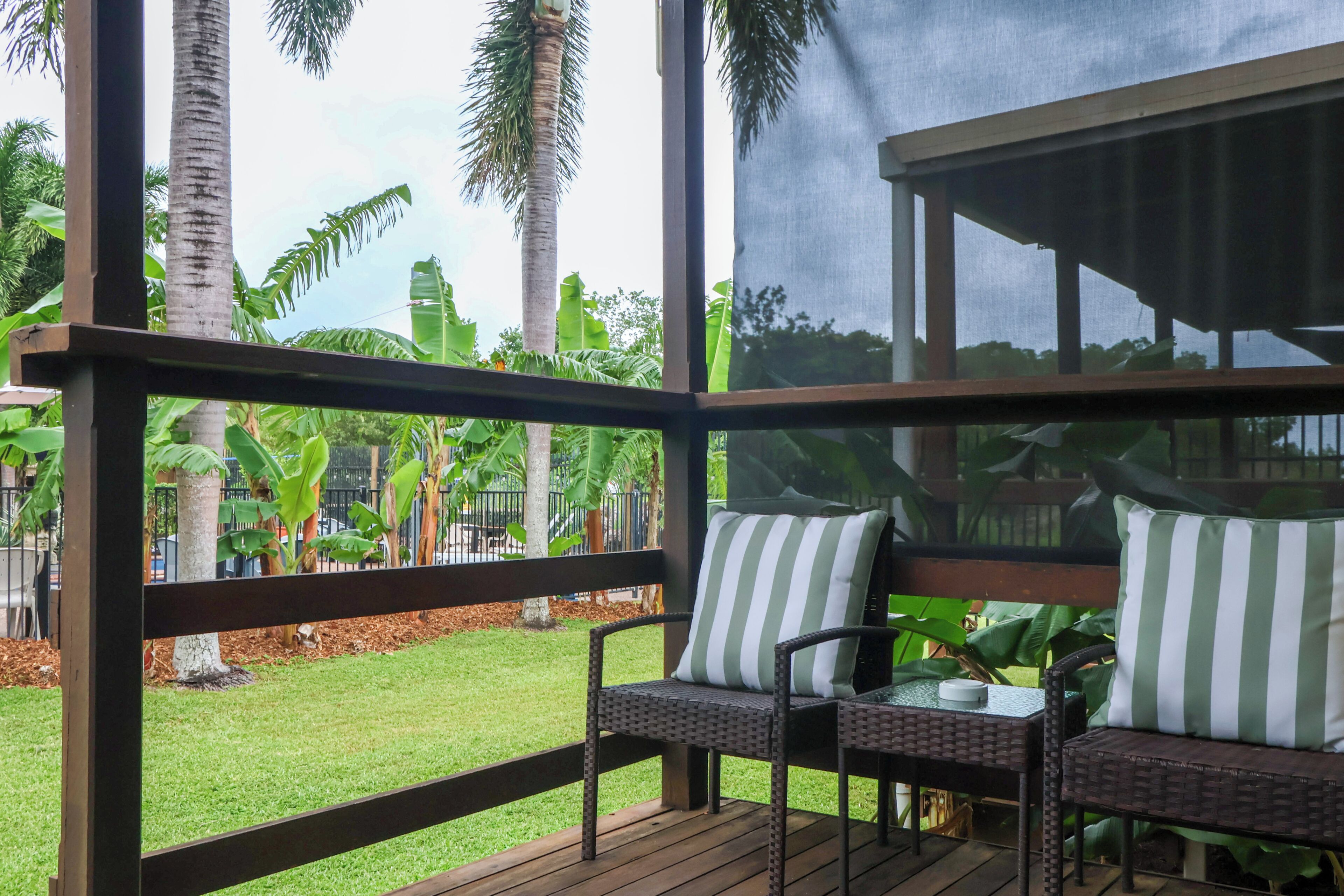 A covered outdoor seating area with wicker furniture and striped cushions, overlooking a well-maintained lawn and palm trees.
