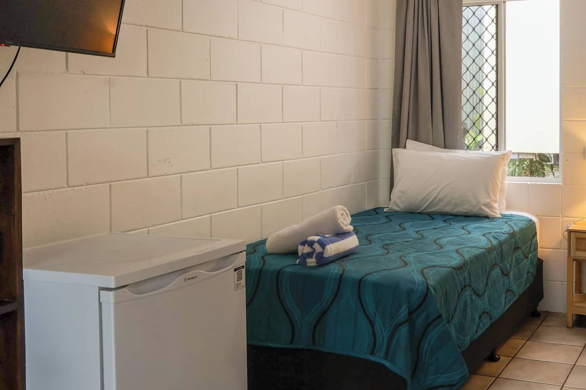 Une petite pièce comprenant un lit, un réfrigérateur blanc, un téléviseur fixé au mur et une fenêtre avec des rideaux.