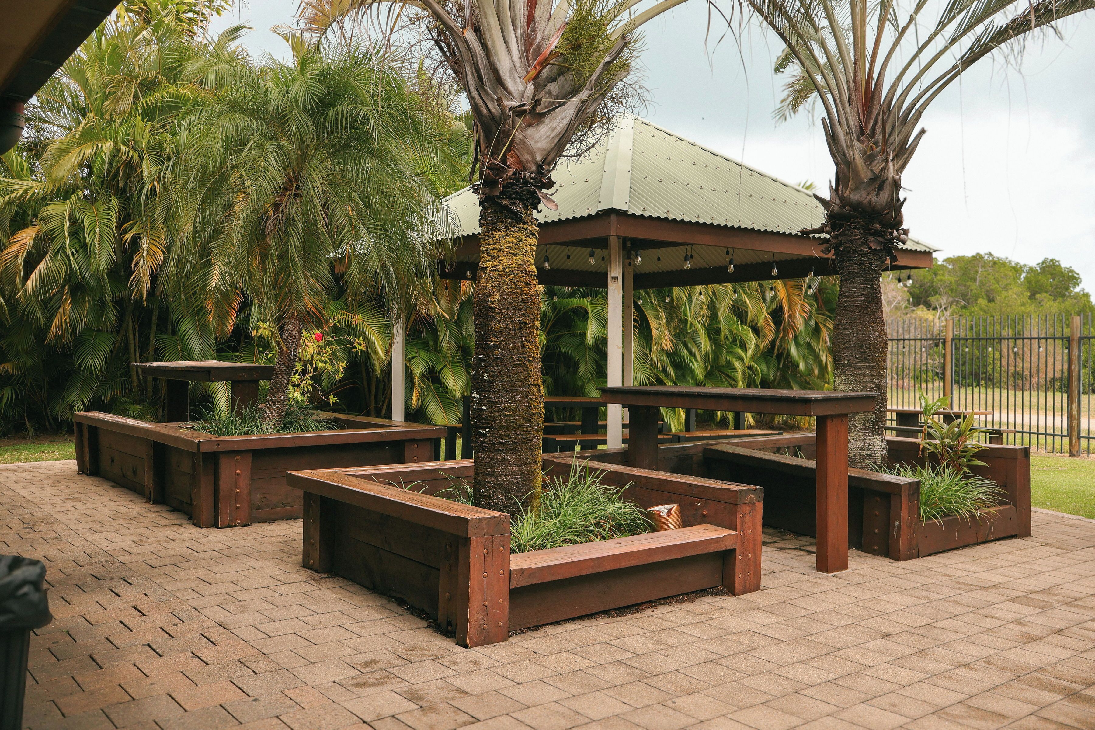 A covered outdoor seating area with benches and planters, surrounded by palm trees.