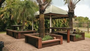 A covered outdoor seating area with benches and planters, surrounded by palm trees.
