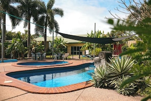 A swimming pool with a brick border, surrounded by palm trees and a shaded area.