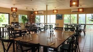 Een restaurant met meerdere tafels, stoelen, hangende lampen en grote ramen met uitzicht op groen.