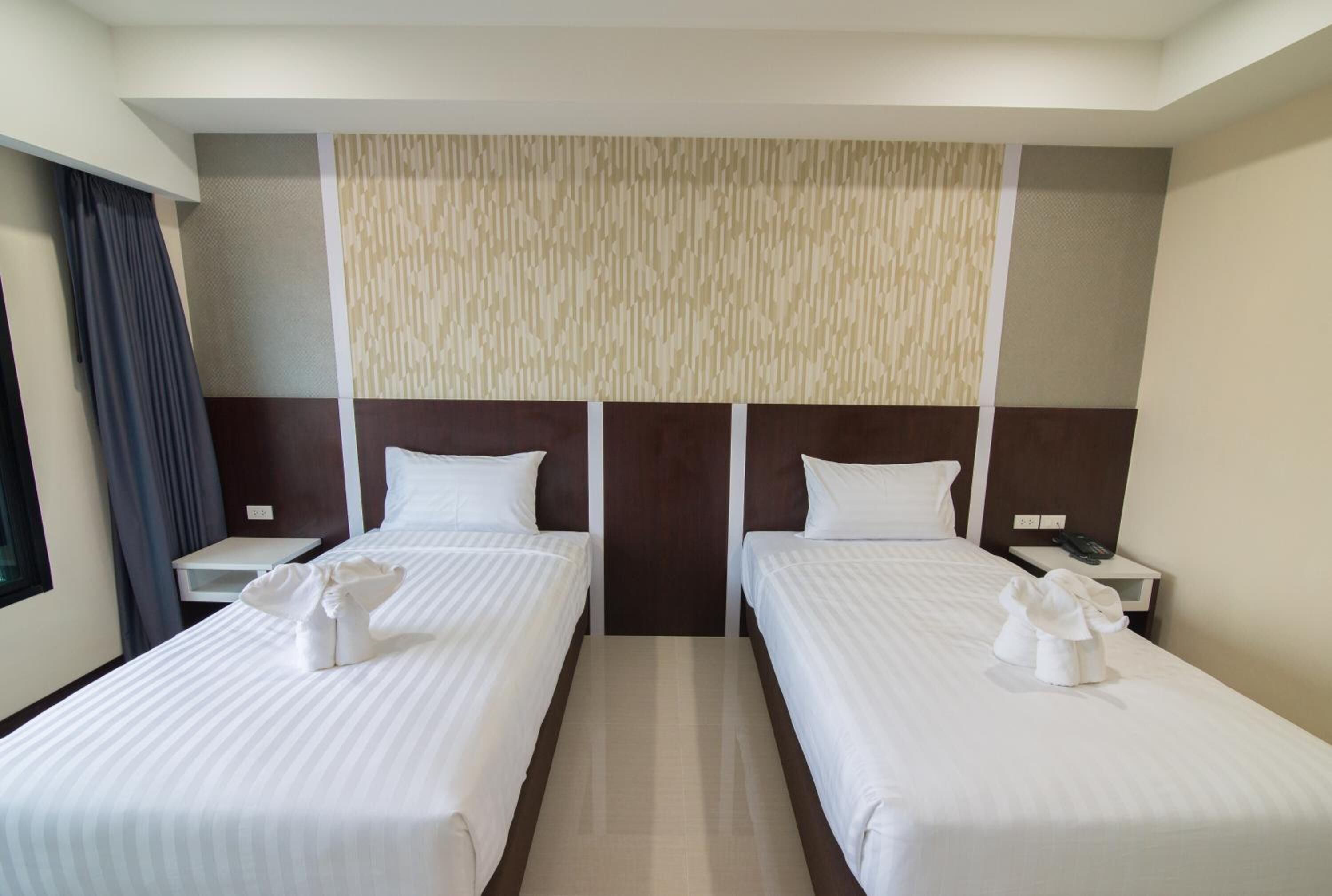 Habitación de hotel con dos camas individuales, un ventanal con cortinas, un panel en la pared y una mesita de noche con teléfono.