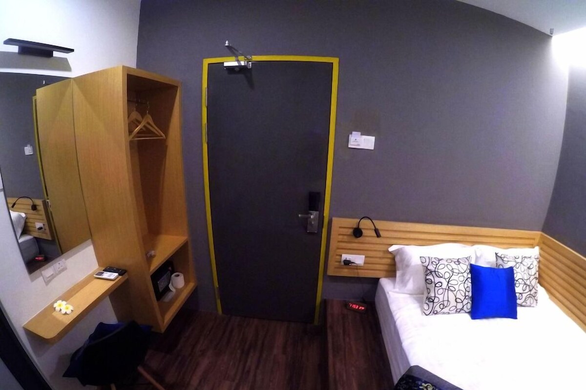 A hotel room with a bed, a wooden wardrobe, a desk with a chair, and a door with a yellow frame.
