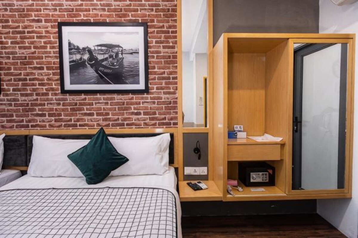 A hotel room with a brick wall, a bed with white bedding and a green pillow, a wooden wardrobe, and a framed picture above the bed.