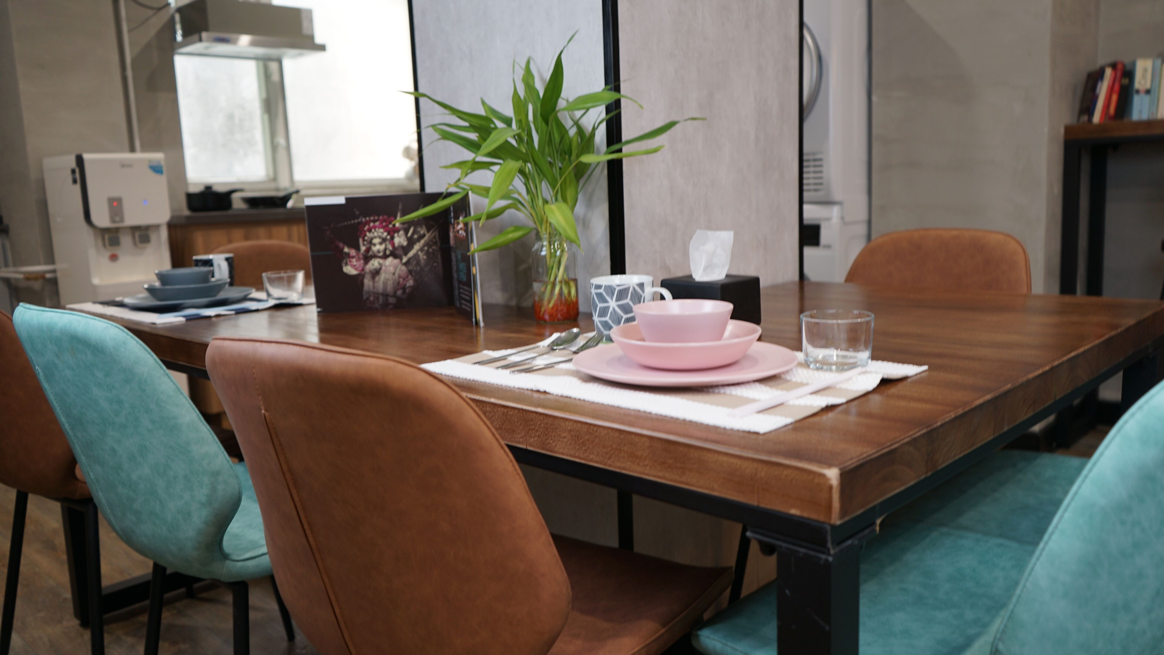 Un comedor con una mesa de madera, sillas azul verdosas y un florero con plantas verdes.