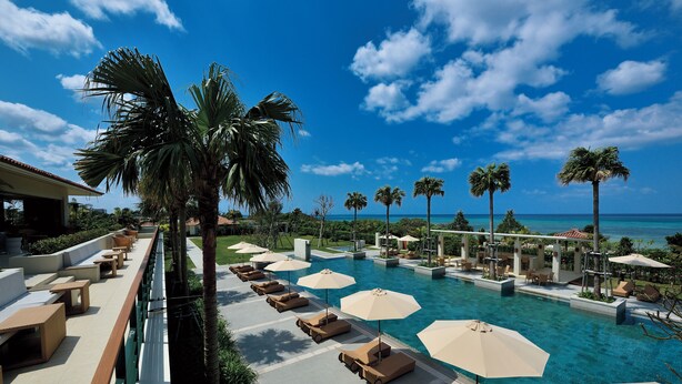 A poolside area with lounge chairs and umbrellas, palm trees, and a view of the ocean. - The Uza Terrace Beach Club Villas (Yomitan)