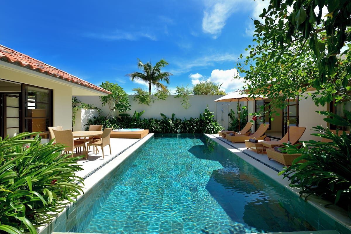 A rectangular swimming pool with clear blue water, surrounded by lounge chairs and a dining area with a table and chairs.