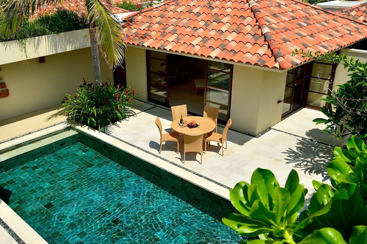 A swimming pool with a tiled deck, surrounded by a patio with wicker furniture and a table.