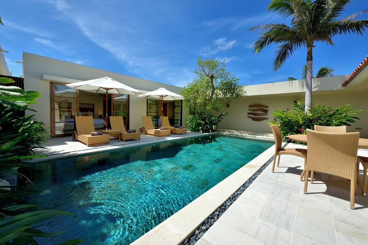 A pool area with lounge chairs and umbrellas, surrounded by a modern building with large windows and a tiled floor.