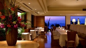 A restaurant with tables and chairs, a large vase of flowers, and a view of the ocean at dusk. - The Uza Terrace Beach Club Villas (Yomitan)