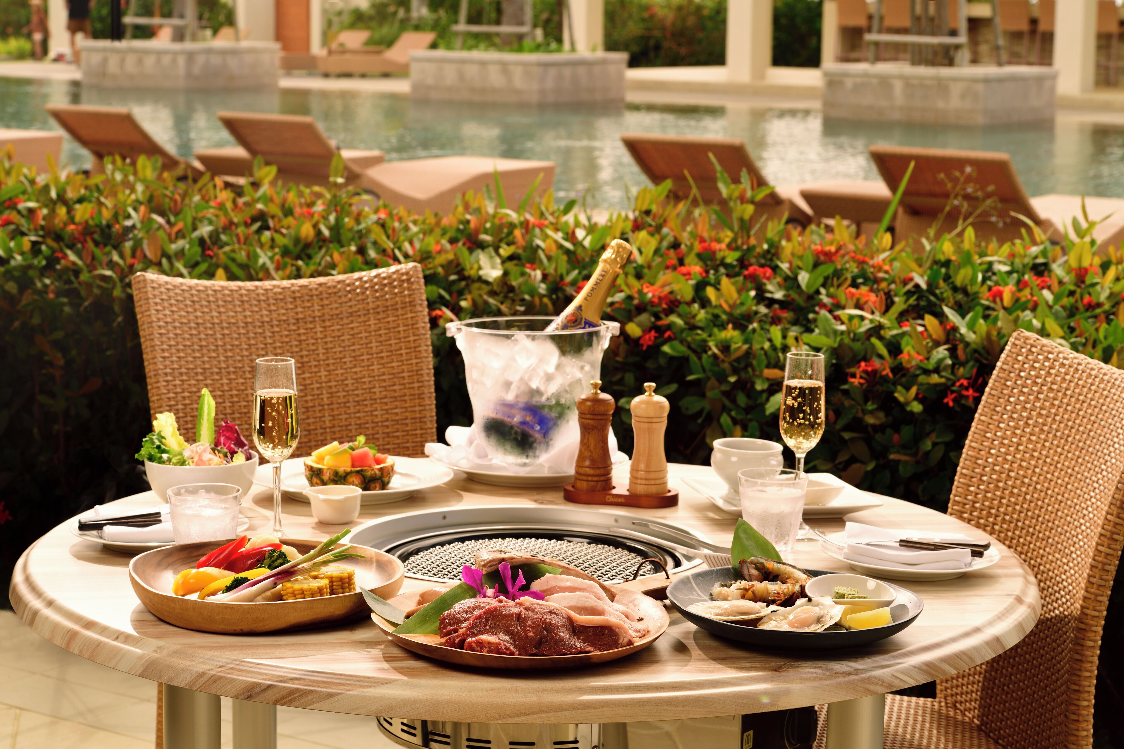Mesa de comedor dispuesta con varios platos, una hornilla caliente y bebidas, rodeada de sillas de mimbre y vista a la piscina.