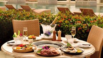 Mesa de comedor dispuesta con varios platos, una hornilla caliente y bebidas, rodeada de sillas de mimbre y vista a la piscina.