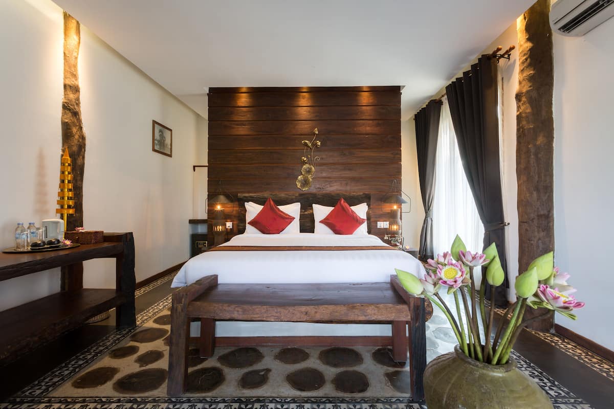 A hotel room with a large bed, wooden headboard, bedside tables, a bench, a vase with flowers, and a patterned rug.