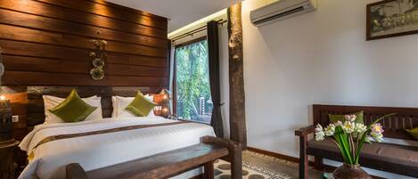 A bedroom with a wooden headboard, a large bed with green pillows, a glass coffee table with a vase of flowers, and a bench.