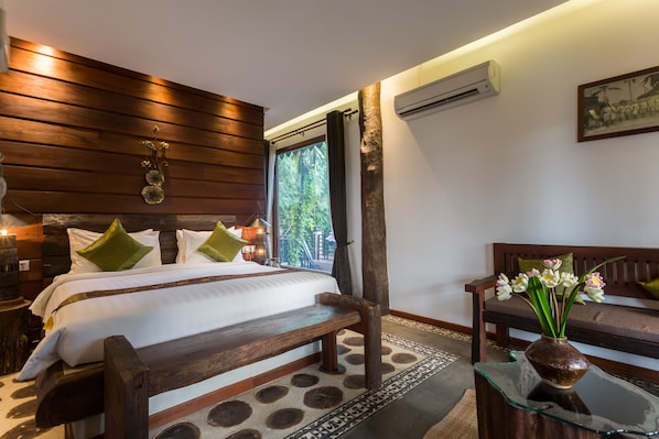 Un dormitorio con cabecera de madera, una cama grande con almohadas verdes, una mesa de centro de vidrio con un florero y un banco.