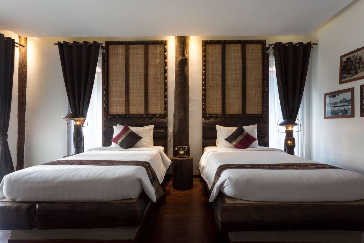 A hotel room with two beds, dark curtains, a small table with a lamp, and framed pictures on the wall.