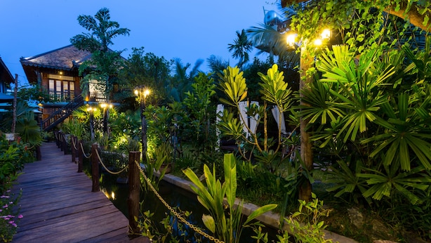 A wooden pathway leading to a building with a thatched roof, surrounded by lush tropical vegetation and a small water feature. - JAVA WOODEN VILLA (Siem Reap)